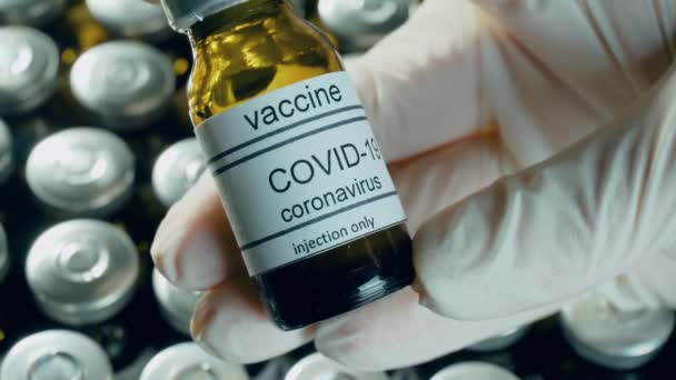 Медичний вірусолог руки в захисних рукавичках, що тримають вакцину проти коронавірусу в флаконі на фармакологічних препаратах на фоні пляшок на тлі пандемії у всьому світі — стокове відео