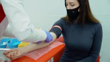 Modern bir klinikte kadın doktor, kadın hasta damarından tüp içine kan testi yaptırıyor, ziyaretçi kliniğe koruyucu yüz maskesi takıyor.