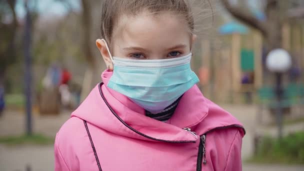 Retrato de niña en máscara médica mira a la cámara de pie en el parque de la ciudad en el soleado día de primavera durante la epidemia mundial de virus — Vídeos de Stock