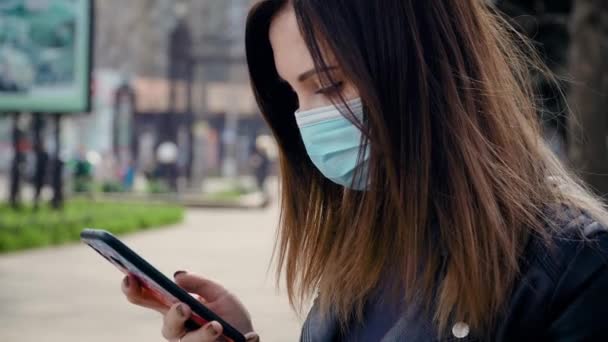 Giovane donna bruna in maschera medica protettiva seduta sulla panchina nel parco utilizza rotoli di telefono naviga su internet e cerca notizie sull'infezione da covid19 durante la pandemia — Video Stock