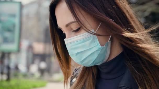 Retrato de una mujer joven con máscara médica mirando hacia abajo de pie en el parque de la ciudad en el día nublado de primavera durante la epidemia mundial de virus y su cabello balanceándose en el viento — Vídeo de stock