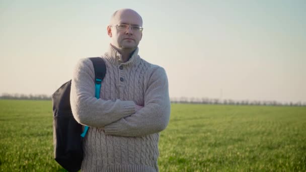 Portrait of free serious man with backpack enjoying nature and standing in large green field at sunset on background of waving in wind crops — Stock Video