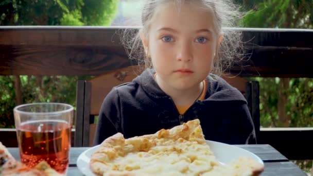 Petite fille mignonne aux cheveux lâches éclairés par le soleil assis à table avec une pizza parfumée coupée en morceaux dans un complexe de luxe tropical — Video