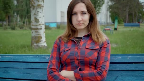 Портрет серьезной женщины средних лет. Женщина в красной рубашке сидит на скамейке на фоне детской площадки в пасмурный день — стоковое видео