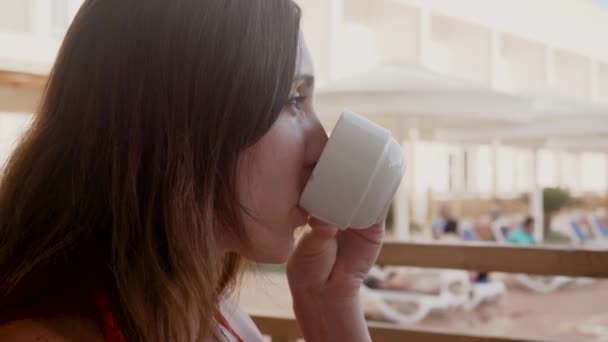 Mujer bebe jugo de la taza y relajarse en la terraza de la habitación del complejo contra de sombrillas y tumbonas. Cuidado de la salud, autenticidad, sentido de equilibrio y calma — Vídeos de Stock