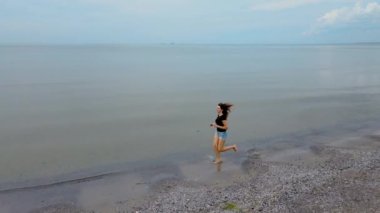 Zayıf bir kadın kumlu sahilde deniz dalgaları boyunca ağır çekimde koşuyor. Güzel bir kadın deniz kıyısında sörf yapıyor. Okyanus kıyısında banyo üstüne kız.