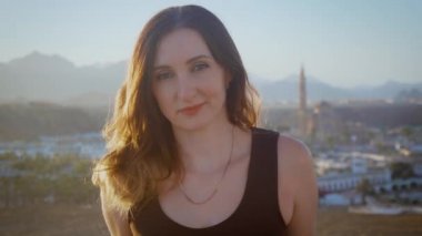 Güzel, esmer, uzun saçlı, gülümseyen bir kadının yakından çekilmiş yüzü. Arap şehri ve camiinin arka planında kameraya bakan muhteşem kızın portresi
