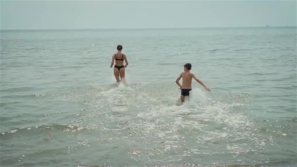 Mother and son run into water. Woman and boy jumping into sea and splashing view from behind slow motion on ocean resort — Vídeos de Stock