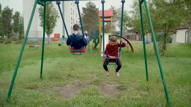 Happy kids are riding on swing. Brother and sister on cloudy spring day on playground. Boy and girl play and develop — Stockvideo