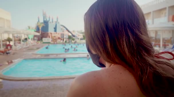 Неузнаваемая женщина в медицинской защитной маске для лица в красном бикини стоит на фоне бассейна в роскошном курорте и смотрит на людей во время всемирной пандемии коронного вируса — стоковое видео