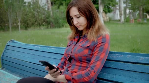 Mensaje de escritura femenina seria de mediana edad en la red social en el teléfono móvil. Mujer con camisa roja se sienta en el banco contra el fondo del patio de recreo en el día nublado — Vídeo de stock