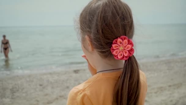 Menina bonito está de pé na praia com doces no pau e olha para silhuetas de sua mãe e irmão saindo da água — Vídeo de Stock