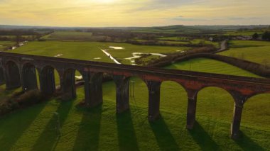 Tarihi demiryolu viyadük, altın akşam gökyüzünün altında yeşil tarlaların üzerinde duruyor. Bahar alacakaranlığında demiryolu köprüsünün havadan bakış açısı. Sıcak günbatımı tonlarıyla yuvarlanan kırlara uzanan görkemli tuğla kemerleri.