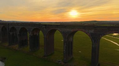 Gün batımında bereketli kırlara uzanan görkemli tuğla viyadük. Tren viyadük gece ışığında tepelerin üzerinden uzanıyor. Geniş demiryolu köprüsü altın gökyüzünün altında canlı yeşil alanlar üzerinde dimdik duruyor.
