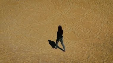 Çıplak ayakla güneşli kumsalda tek başına yürüyen bir kadın. Dişi, dağılmış ayak izleriyle dokulu altın zemin üzerinde yavaşça hareket ediyor. Terkedilmiş sahilde sakince gezinen kadın yumuşak kumlarda izler bırakıyor.