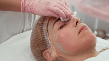 Modern kozmetoloji salonunda yüz temizleme ameliyatı olan bir kadın ameliyat sırasında sakince yalan söylüyor. Güzellik uzmanı maske kalıntılarını çıkarmak için nazikçe yüzünü siliyor ve cildini tazeliyor. Profesyonel cilt bakımını seven hasta yenilenmeye ve