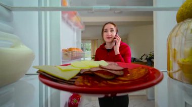 Kadın buzdolabını açıyor, cep telefonuyla konuşuyor ve yanlışlıkla içine yemek yerine telefon koyuyor buzdolabının içini görüyor. Genç bayan dikkati dağılmış ve eğlenmiş görünüyor. Telefonla konuşurken elinde sosisli ve peynirli kırmızı tabak tutan kadın.