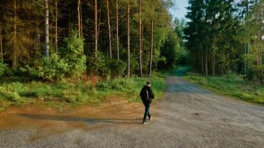Güneşli bir havada orman kavşağında yürüyen bir adam. Explorer açık gökyüzünün altından geçerek ormanlık alandan geçiyor. Ağaçlar ve ışıkla çevrili çakıl yolu boyunca gezinen sırt çantalı.