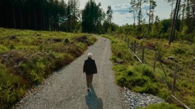 Yaşlı kadın orman yolunda yürüyor ve öğleden sonra güneşinin tadını çıkarıyor. Emekliler çakıllı yolda rahatça dolaşır. Yaşlı kadın ormanlık alanda yürüyor. Huzurlu bir çevreyi kucaklıyor.