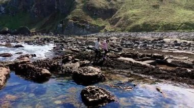 Anne ve kızı okyanus dalgaları yakınında kayalık sahilde yürüyor. Kadın ve kız, dalgalanan denizin kıyısındaki taşlarda geziniyorlar. Ebeveynler ve çocuklar deniz kenarındaki kayalarda sörf yapmaya gidiyorlar.