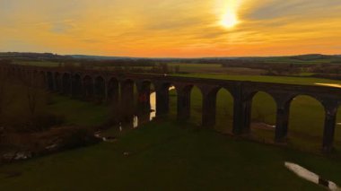 Altın gökyüzünün altındaki yeşil vadiyi kaplayan uzun demiryolu viyameti. Solan güneşin altında parlayan tarihi taş viyadük. Sonsuz tuğla köprü sıcak alacakaranlık renklerinde uçsuz bucaksız kırsalda yaylanıyor.