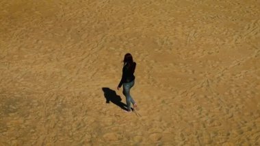 Güneşli bir günde çıplak ayakla kuru kumda yürüyen bir kadın. Dişi, uzun gölgeli, boş altın plajda yavaşça geziniyor. Parlak gökyüzünün altındaki sıcak kıyı bölgesinde tek başına hareket eden bir kadın.