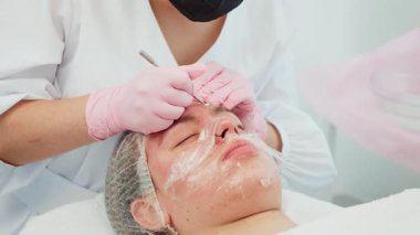 Rahatlamış yüzüyle tedavi yatağında yatan bir kadın, eldivenli bir güzellik uzmanı akne temizleme ameliyatı yapıyor. Kadın yüz tazeleme ve gözenek temizleme tedavisi görüyor. Sakin klinik ortamında profesyonel cilt bakımı gören kadın