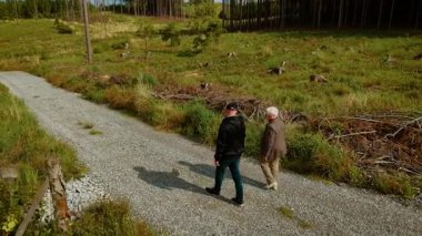 Oğul, güneşli havada orman çakıl yolunda yaşlı annenin yanında yürüyor. Açık gökyüzünün altındaki yeşil kırsal yolda yetişkin bir çocuğun yanında gezinen bir kadın. Erkek ve yaşlı kadın, çimen ve ağaçlarla çerçevelenmiş açık ormanlık yolda birlikte yürüyüş yapıyor.