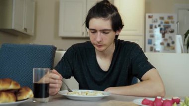 Genç adam sakin bir ifadeyle mutfakta otururken köfteler tabakta. İçerideki kişi yemeğe devam etmeden önce dikkatli bir şekilde bakıyor. Masadaki adam, geleneksel yemekleri sessiz kafayla yemeye hazırlanıyor.