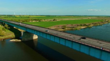 Araba ve kargo minibüsü arka planda rüzgar türbinleriyle köprüden karşıya geçiyor. Araçlar yeşil tarlaların yakınındaki su yolu boyunca ilerliyorlar. Yolcu otomobili ve teslimat minibüsü türbinlerle akan nehrin yukarısında viyadük üzerinde seyahat ediyor.