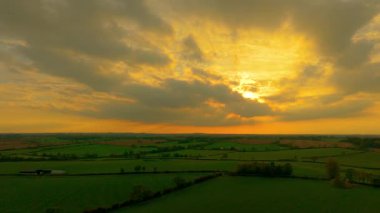 Akşam vakti yeşil tarım arazisi üzerinde bulutların arasından gün ışığı doğuyor. Altın kirişler tarlalara dokunarak sıcak bir parıltı yayıyor. Kırsal kesimi aydınlatan gün batımı huzurlu tarım manzarası yaratıyor