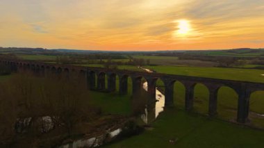 Gün batımında huzurlu kırsal kesimde manzaralı bir viyadük. Resimli demiryolu köprüsü alacakaranlıkta ele geçirildi. Geniş tarihi yapı, sıcak akşam gökyüzü altında yuvarlanan yeşil arazi boyunca uzanıyor.