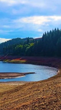 Kurumuş rezervuar kıyı şeridi ve sık ormanlarla çevrili sular. Düşük su seviyesi mavi gölün yanındaki kayalık zemini gösteriyor. Küçülen rezervuar ve açıkta kalan toprak, devam eden kuraklığı gösteriyor.