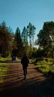 Güneşli bir havada park halindeki arabaya doğru yürüyen bir yürüyüşçü. Sırt çantalı gezgin, aydınlık bir günde ağaçlık yolda yürüyor, dikey oryantasyon. Gezgin, açık mavi gökyüzünün altında orman yolunda ilerliyor.
