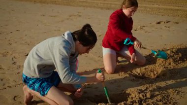 Çocuklar renkli küreklerle kumda derin çukurlar kazıyor, birlikte gülümsüyorlar. Erkek ve kız kardeşler kumsal inşaatında çalışıyor, güneşli günün tadını çıkarıyorlar. Gençler neşeli kum projesi yapıyor.