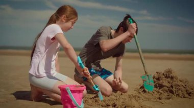 Kız küçük kürekle kum kazarken oğlan büyük kürekle çalışıp eğleniyor. Çocuklar kova ve aletlerle kumsal inşaatı yapıyor, kumsalda gülümsüyor. Erkek ve kız kardeş birlikte kum projesi yapıyor, güneşin altında eğleniyorlar.