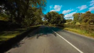 Uzun ağaçlarla gölgelenmiş ve çimenli tarlalarla çevrili kırsal yolda ilerliyoruz. Yeşillik ve elektrik direkleriyle kaplı kırsal otoban boyunca ilerliyoruz. Manzaralı asfalt yolda ilerlerken ormandan geçer ve açılır.