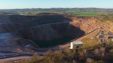 Derin kazılar ve dipte su toplanması etrafında çalışan ağır makinelerle açık maden ocağı arazisi. Teraslı kaya duvarları ve taşıma yolları boyunca çalışan endüstriyel ekipmanlarla büyük ölçekli taş ocağı çıkarma. Mineralin hava görüntüsü