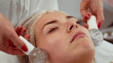 Kozmetik uzmanı spa salonunda kürelerle yüz masajı yapıyor, rahatlama ve cilt tazeleme üzerine çalışıyor. Kadın huzur içinde uzanıyor, rahatlatıcı ve serinletici terapinin tadını çıkarıyor. Yenilenmiş ve ışıl ışıl cilt tedavisi gören hasta