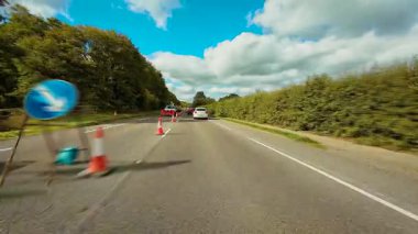 Trafik konileri ve tamir ekipmanlarıyla yol inşaatı bölgesinde araba sürüyor. Otomobil otoyol boyunca ağaçlar ve bulutlu gökyüzü ile çevrili bir şekilde hareket ediyor. SUV uyarı işaretleriyle yol çalışması alanından geçiyor.