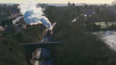 Havadan bakıldığında buharlı lokomotif demiryolu boyunca ilerliyor ve beyaz buhar ve duman yayıyor. Antika demiryolu motoru banliyö arazisinde hareket ederek yükselen buhar bulutları yayıyor. Tarihi buhar treni demiryolu koridorunda ilerliyor.