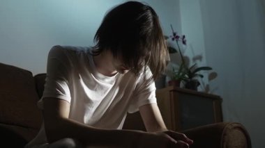 Genç adam düşünceli bir şekilde yalnız başına karanlık odada oturuyor. Duygusal bir ağırlık hissediyor. İç gerilim ve üzüntüyle yüzleşen kişi sessiz bir yansımayla öne doğru eğiliyor. Genç, loş uzayda tek başına dinleniyor derin düşünceler ve psikolojik deneyimler yaşıyor.