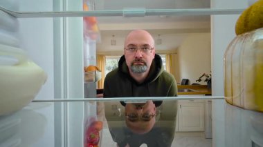 Buzdolabını açan ve içi boş boş bakan, ilgisiz bir yüz ifadesi olan bir adam, buzdolabının içini görüyor. Erkek, ilgisiz ve pasif hissederken içeriğini kontrol ediyor. Adam hareketsiz duruyor ve duygusuz ve yorgunken boş rafları inceliyor.