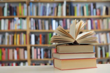 Fotoğraf, hem tanıdık hem de nazikçe idealize edilmiş gibi hissettiren sessiz bir sahne sunuyor: üzerinde açık bir kitap bulunan, pürüzsüz, soluk bir yüzey üzerinde dinlenen küçük bir kitap yığını, sayfaları çırpınışların ortasında kalmış.