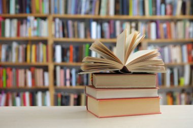 Fotoğraf, hem tanıdık hem de nazikçe idealize edilmiş gibi hissettiren sessiz bir sahne sunuyor: üzerinde açık bir kitap bulunan, pürüzsüz, soluk bir yüzey üzerinde dinlenen küçük bir kitap yığını, sayfaları çırpınışların ortasında kalmış.