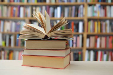 Fotoğraf, hem tanıdık hem de nazikçe idealize edilmiş gibi hissettiren sessiz bir sahne sunuyor: üzerinde açık bir kitap bulunan, pürüzsüz, soluk bir yüzey üzerinde dinlenen küçük bir kitap yığını, sayfaları çırpınışların ortasında kalmış.