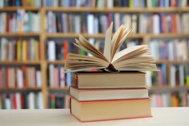 Fotoğraf, hem tanıdık hem de nazikçe idealize edilmiş gibi hissettiren sessiz bir sahne sunuyor: üzerinde açık bir kitap bulunan, pürüzsüz, soluk bir yüzey üzerinde dinlenen küçük bir kitap yığını, sayfaları çırpınışların ortasında kalmış.