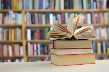 Fotoğraf, hem tanıdık hem de nazikçe idealize edilmiş gibi hissettiren sessiz bir sahne sunuyor: üzerinde açık bir kitap bulunan, pürüzsüz, soluk bir yüzey üzerinde dinlenen küçük bir kitap yığını, sayfaları çırpınışların ortasında kalmış.