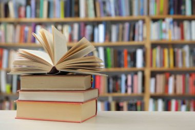 Fotoğraf, hem tanıdık hem de nazikçe idealize edilmiş gibi hissettiren sessiz bir sahne sunuyor: üzerinde açık bir kitap bulunan, pürüzsüz, soluk bir yüzey üzerinde dinlenen küçük bir kitap yığını, sayfaları çırpınışların ortasında kalmış.