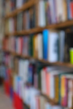 Görüntü, kitaplarla dolu geniş bir raf duvarını gösteriyor, ancak tüm sahne bilerek odak dışı kalmış, yumuşak ve bulanık bir etki yaratıyor. Raflar açık renkli ahşaptan yapılmış gibi görünüyor.,
